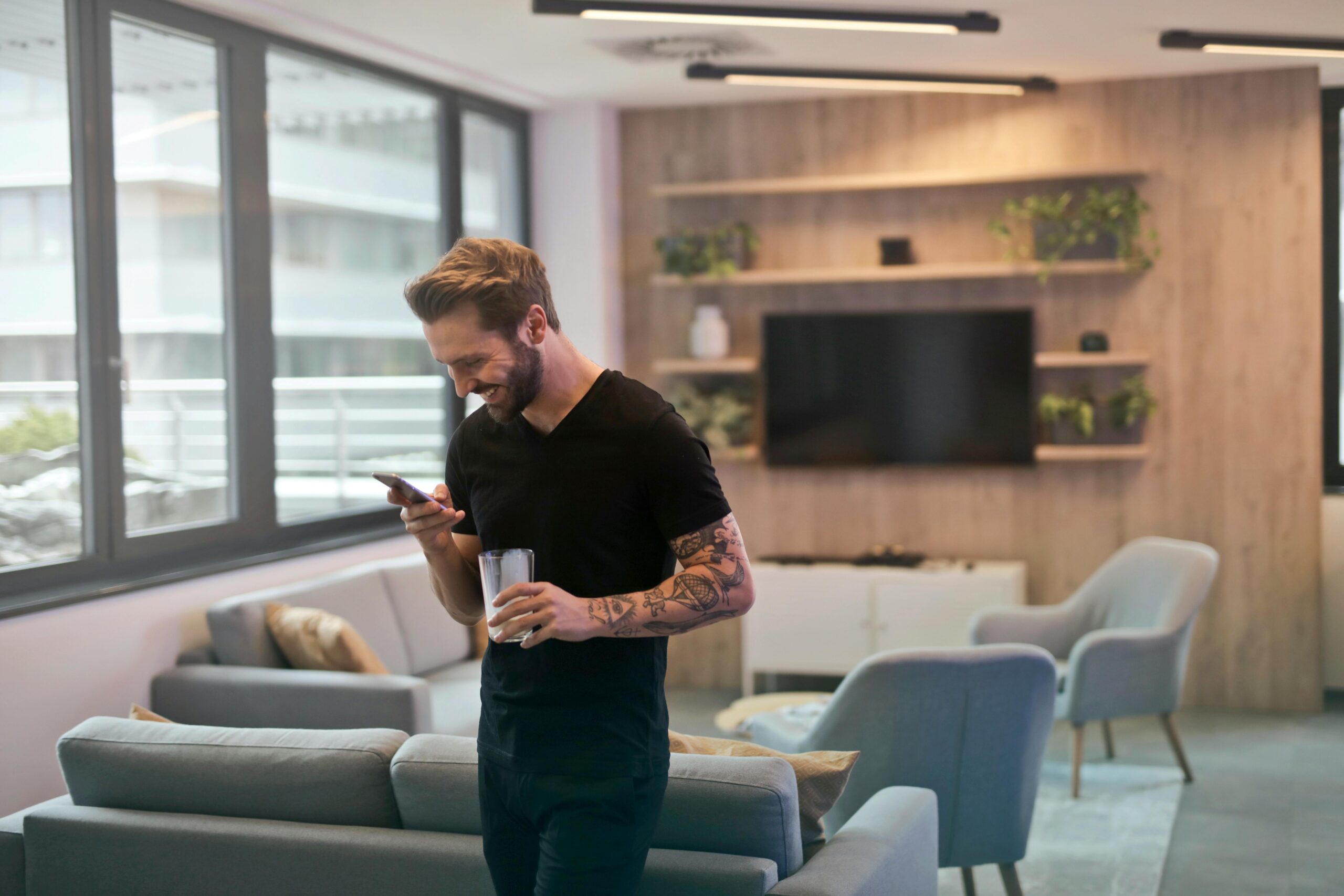 Casual man with tattoos laughing while texting on smartphone in modern office lounge.